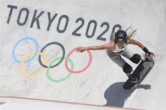 Sky Brown, a young girl, is skating in the bowl at the 2020 Olympic Games. Behind her a mural shows the Olympic Rings and the words