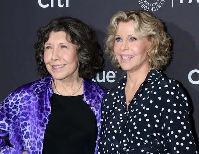 two older women — one brunette on the left wearing a purple leopard print jacket over a black tshirt, one blonde wearing a button up navy shirt with white polka dots — smile at an off-screen camera