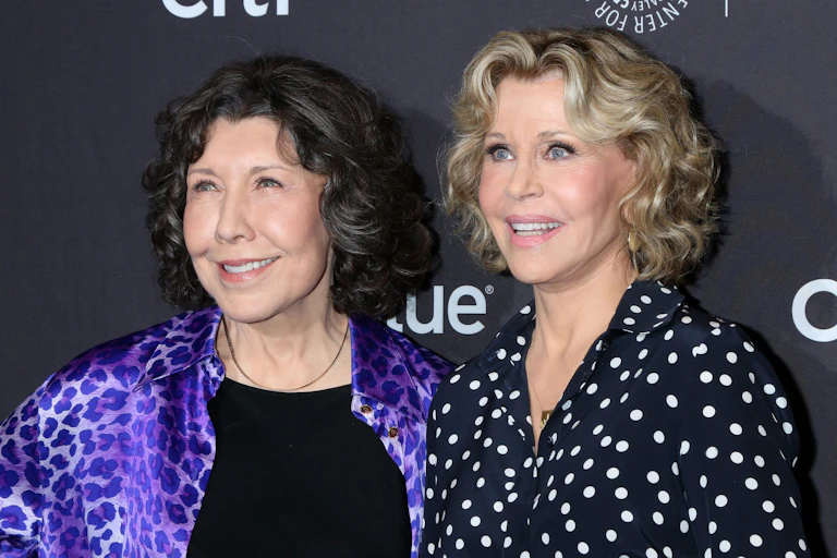 two older women — one brunette on the left wearing a purple leopard print jacket over a black tshirt, one blonde wearing a button up navy shirt with white polka dots — smile at an off-screen camera