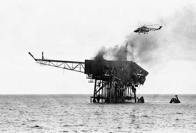 Black and white archive photo of a helicopter flying over the wreckage of a burned oil rig