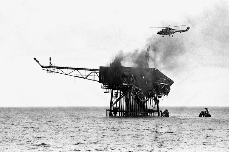 Black and white archive photo of a helicopter flying over the wreckage of a burned oil rig