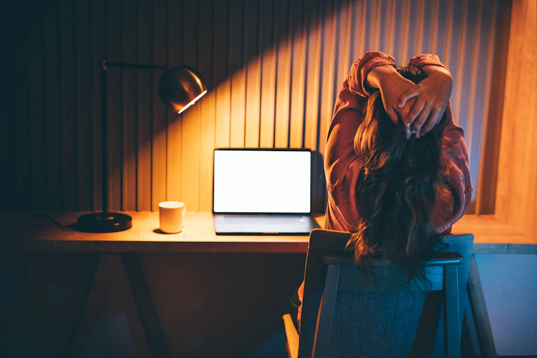 A woman with her hands on the back of her head sits at a computer, seemingly frustrated.