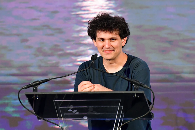 A man with messy hair wearing a t-shirt speaks at a podium