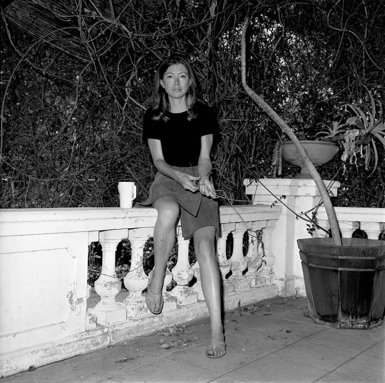 Black and white portrait. Joan Didion sits on a low wall in a knee-length skirt, long hair down by her shoulders. A mug is perched to her left.