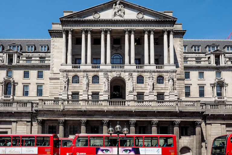 Red buses lined up in front of the Bank of England, people, blue sky.