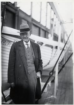 A black and white photo shows a man in a suit and long coat standing in front of a boat.