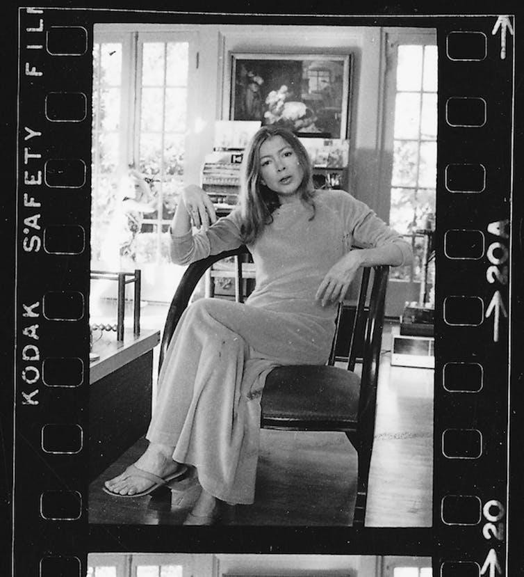 Black and white portrait. Joan sits in her home in an ankle-length dress, looking to camera, one leg crossed over the other.