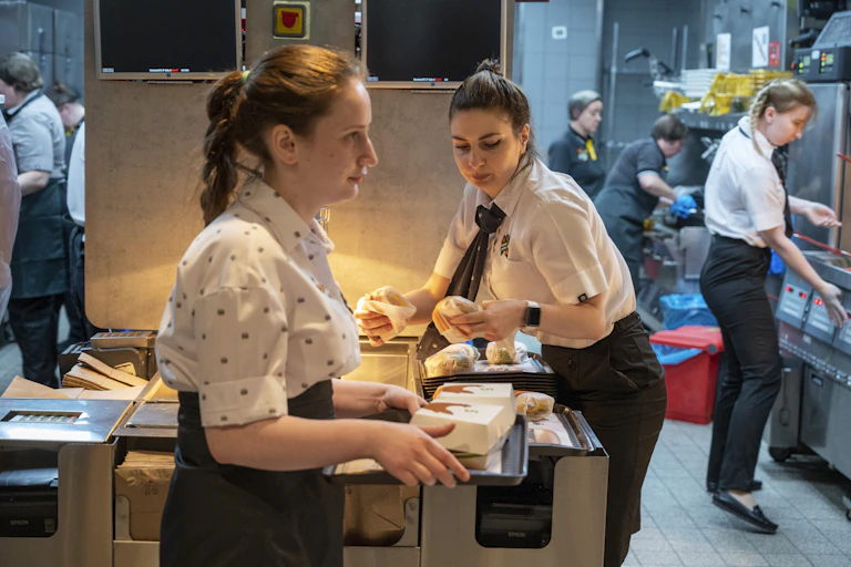 Women with their hair tied back package and transport hamburgers and other fast-food items in a commercial kitchen.