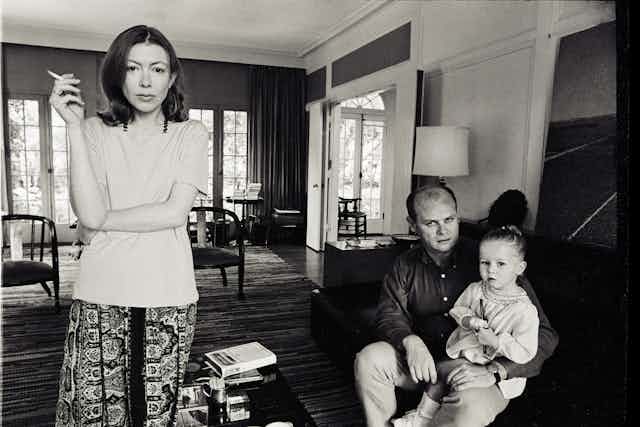 Joan smokes at the left of the frame, looking directly to camera. On the right, her husband sits with their young daughter sat on his lap
