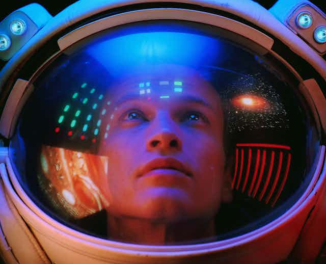 A young man wearing a space helmet peers upward as computers and images of stars and planets reflect in the face shield of the helmet.