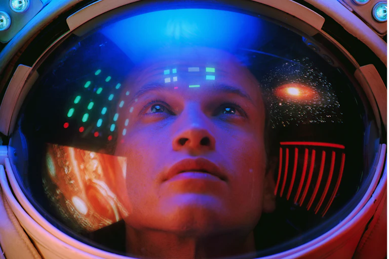 A young man wearing a space helmet peers upward as computers and images of stars and planets reflect in the face shield of the helmet.