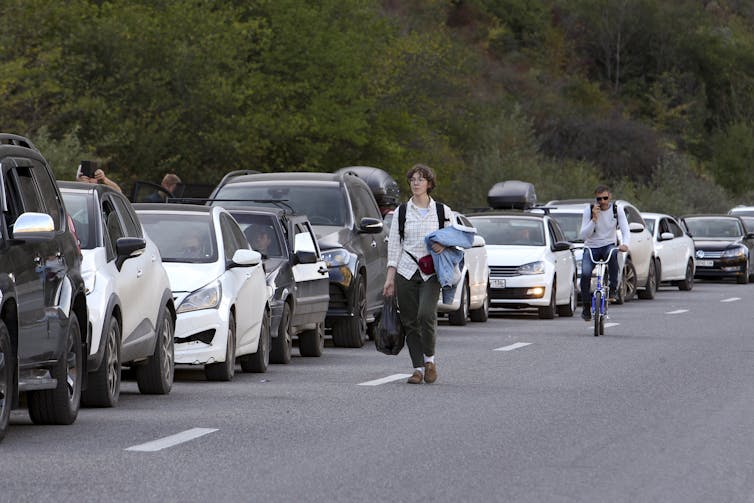 A young man walks as another rides his bicycle beside a long line of cars on a divided roadway.