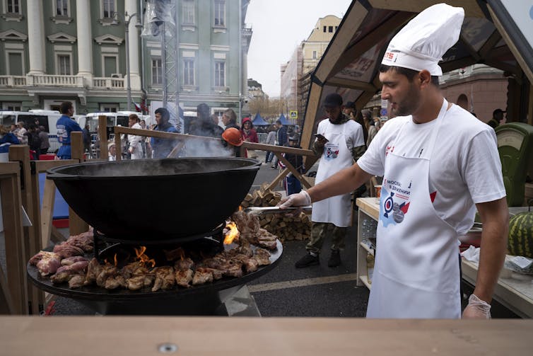 Men in white, one wearing a white chef's hat, barbecue in a city square.