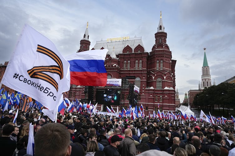 Hundreds of people gather in front of an ornate red brick building waving various flags.