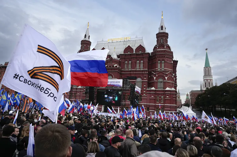 Hundreds of people gather in front of an ornate red brick building waving various flags.