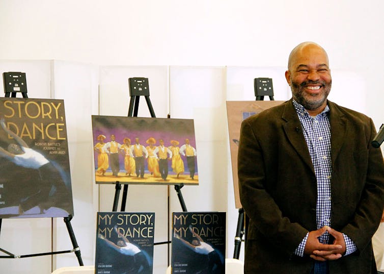 A man in a suit jacket stands grinning in front of a few easels with copies of a book called 'My Story, My Dance.'