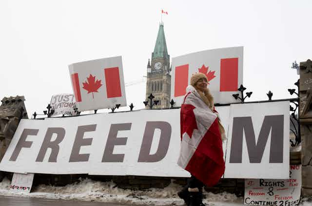 A woman wrapped in a flag walks past a large sign reading Freedom with the Peace Tower in the background.