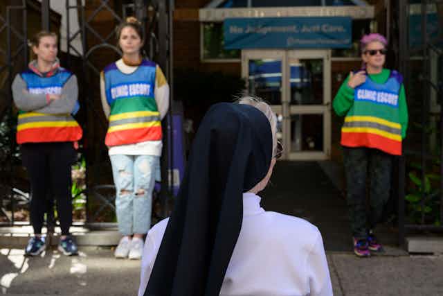 A woman with a black head covering, appearing to be a nun, stands in front of a building with three people wearing rainbow vests bearing the words 'clinic escort'
