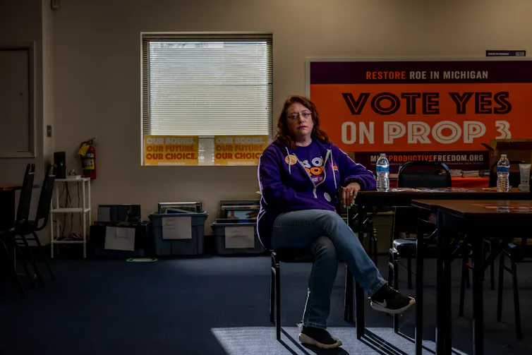 A middle aged woman with brown hair sits on a chair in a room with signs bearing such messages as 'Our bodies, our future, our choice.'