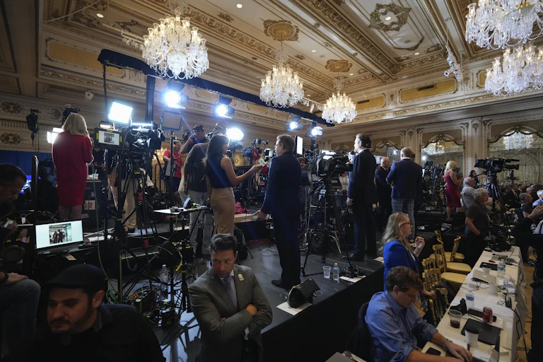People stand in front of cameras and lights, and sit at tables with laptop computers open in front of them.