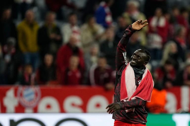 A man in a football tracksuit with a towel around his neck smiles broadly as he waves to a crowd.