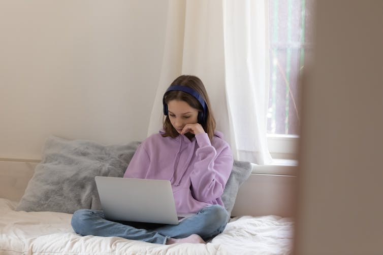 A teenage girl sits on her bed using her laptop.