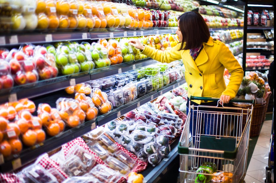 A woman in a yellow coat looking at fruit prices in a shop, pushing a trolley.