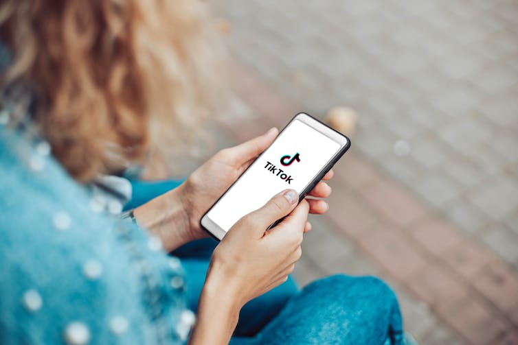 A young woman is photographed from behind opening the TikTok app on her smartphone.