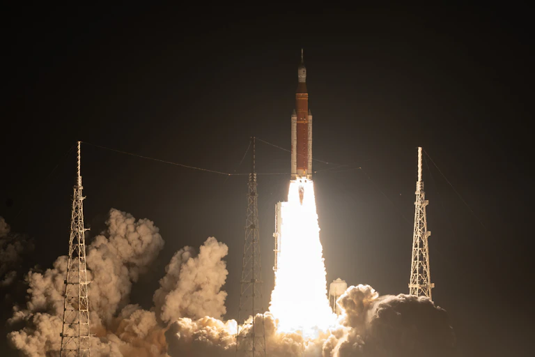 a rocket with flames shooting out of the bottom lifting off the ground as smoke billows around three pylons