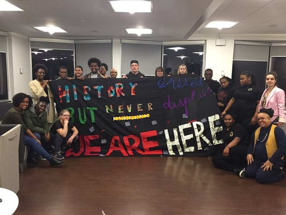 A group of people hold a sign that reads: history erased but never displaced. We are here.