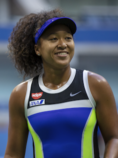 Tennis player Naomi Osaka smiles with her hair in a high ponytail, wearing a blue visor hat