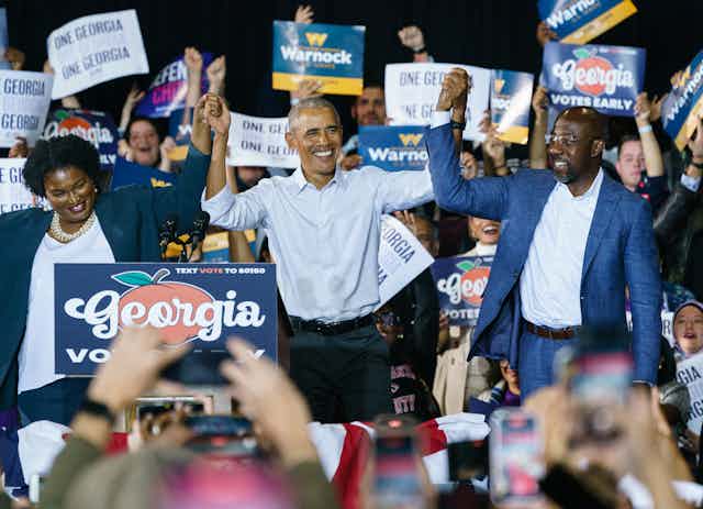 A black woman and two black men are holding up their hands while onlookers applaud and take photographs with their cell phones.