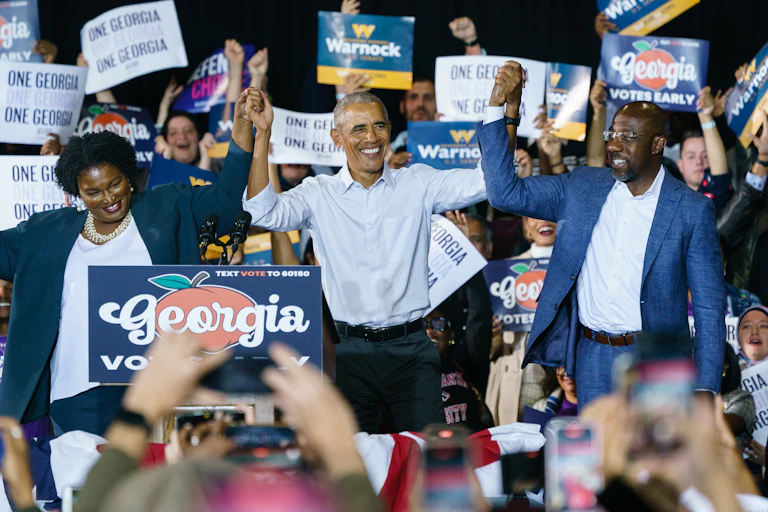 A black woman and two black men are holding up their hands while onlookers applaud and take photographs with their cell phones.