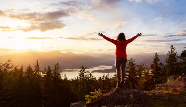 A woman stands on a rock with her arms raised looking out over a scenic remote vista.