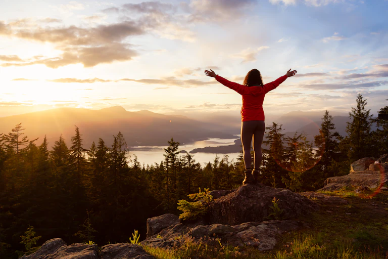 A woman stands on a rock with her arms raised looking out over a scenic remote vista.