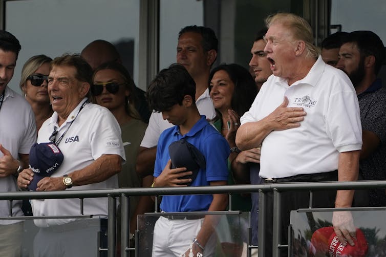 A rotund man in a white T-shirt holds his hand on his heart as he sings.