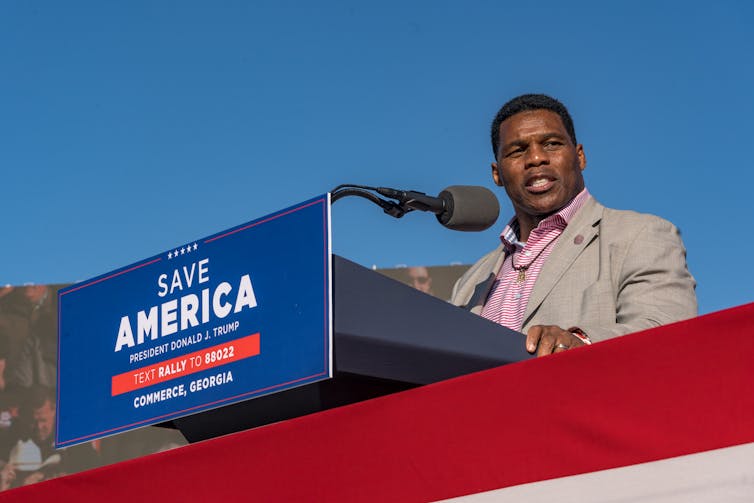 A black man in a suit stands behind a microphone and gives a campaign speech.