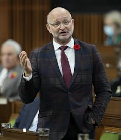 A bald man wearing a suit and glasses speaking in parliament.