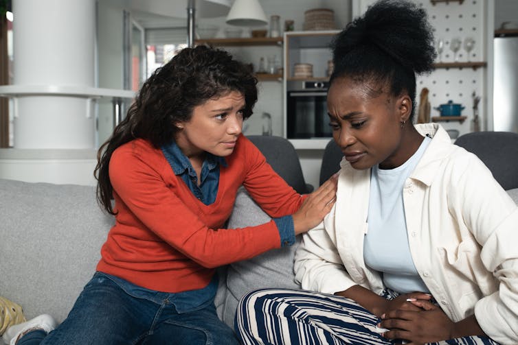 Two women sit on a couch. One is comforting the other.