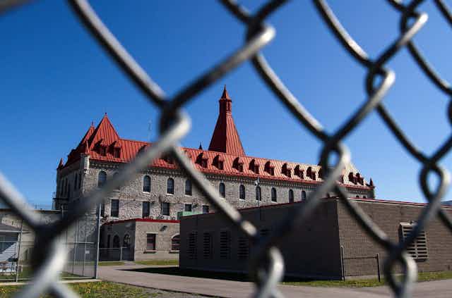 A prison with a red peaked roof is seen through a chain-link fence.