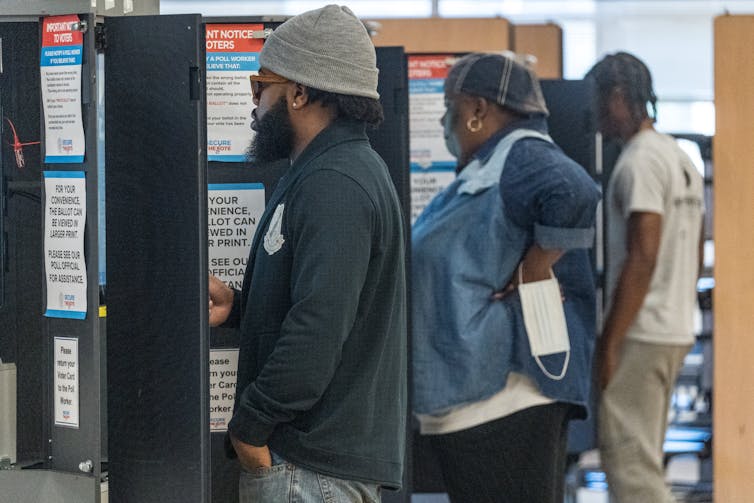 Dos hombres negros y una mujer negra están de pie en las cabinas de votación mientras emiten sus votos.