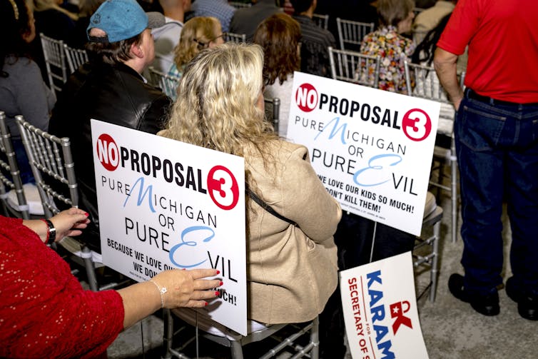 Rows of people sit in back-to-back chairs and hold signs that read 'No Proposal 3 - Pure Michigan or Pure Evil'.
