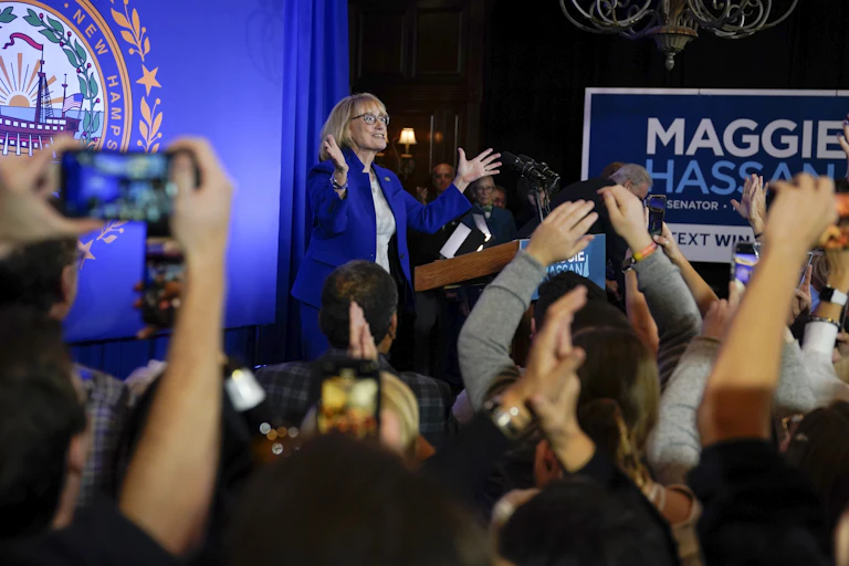 A blond woman in a blue jacket standing on a stage with arms outstretched and many people in front of her.