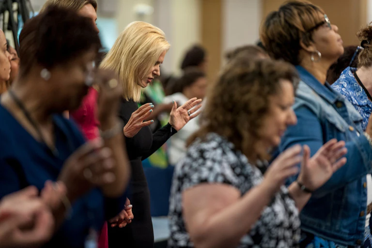 A handful of women pray while standing with their hands raised and their eyes closed.