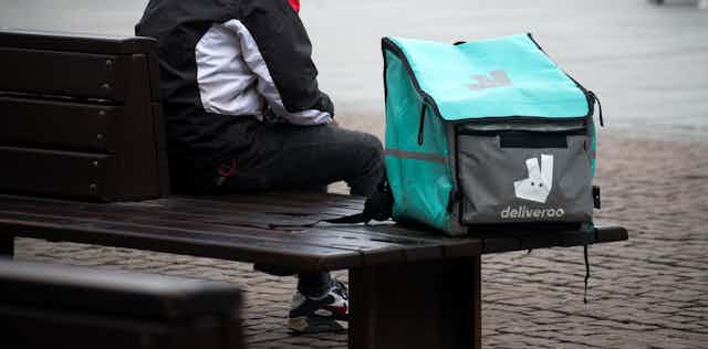 Deliveroo worker sitting on street bench.