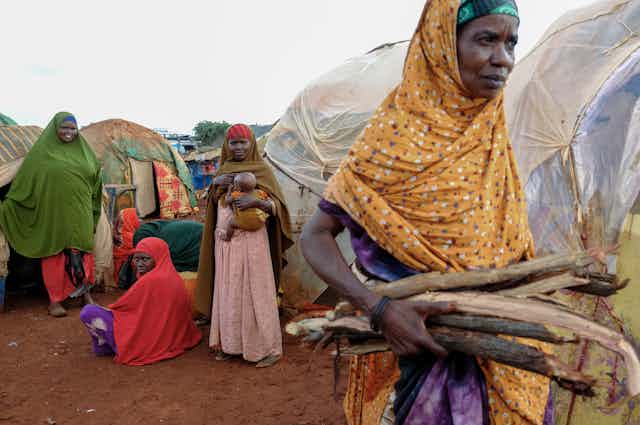 Women in a camp, one carries firewood
