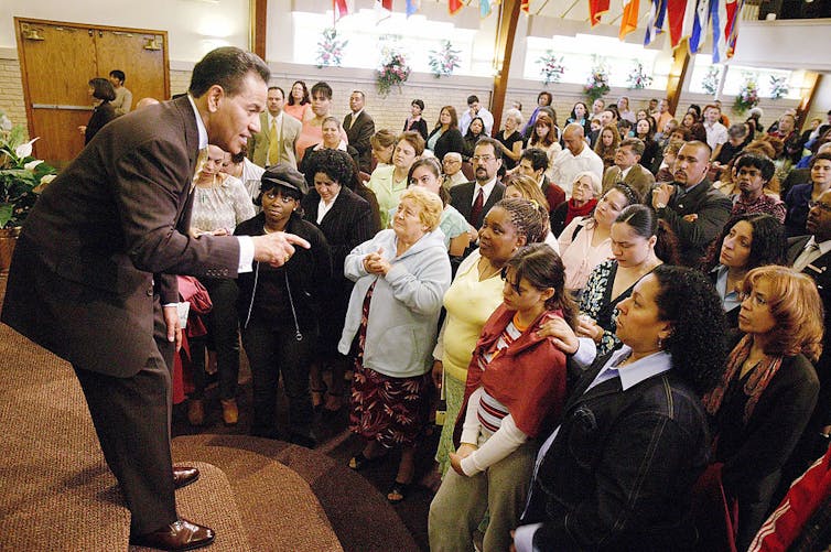 A man in a suit stands on a platform, talking to a large crowd in a room with flags lined up on one wall.