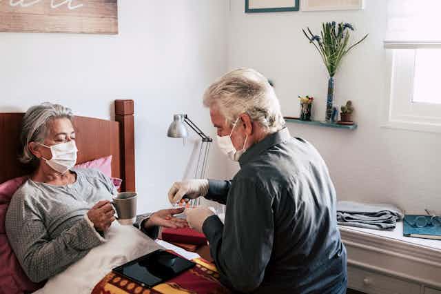 A man gives tablets to his wife who is in bed. Both are wearing masks.