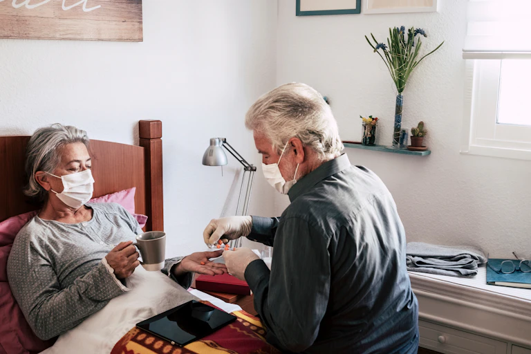 A man gives tablets to his wife who is in bed. Both are wearing masks.