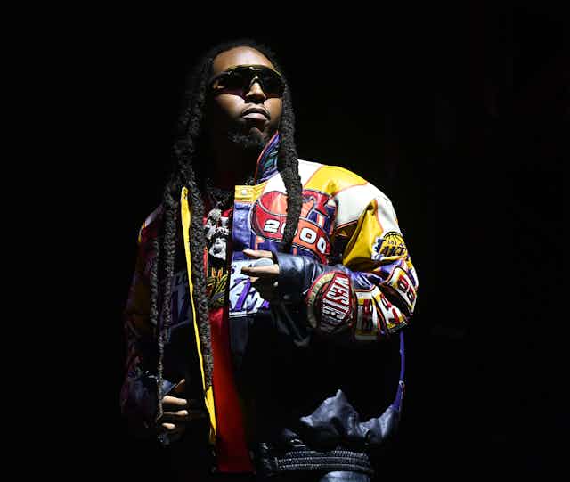 A young black man wearing dark sunglasses stands in the light on a dark stage.